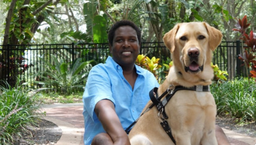 Man sits with yellow goldador in front and both look at the camera and smile