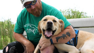 Man sits on a bench with yellow lab service dog lays on the bench and looks at the camera