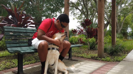 A teenage boy in a red shirt seated on a bench hugs his yellow lab guide dog