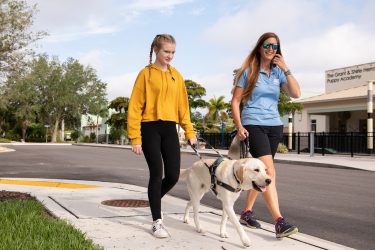 A young teen walks with a guide dog down the sidewalk along with a guide dog trainer