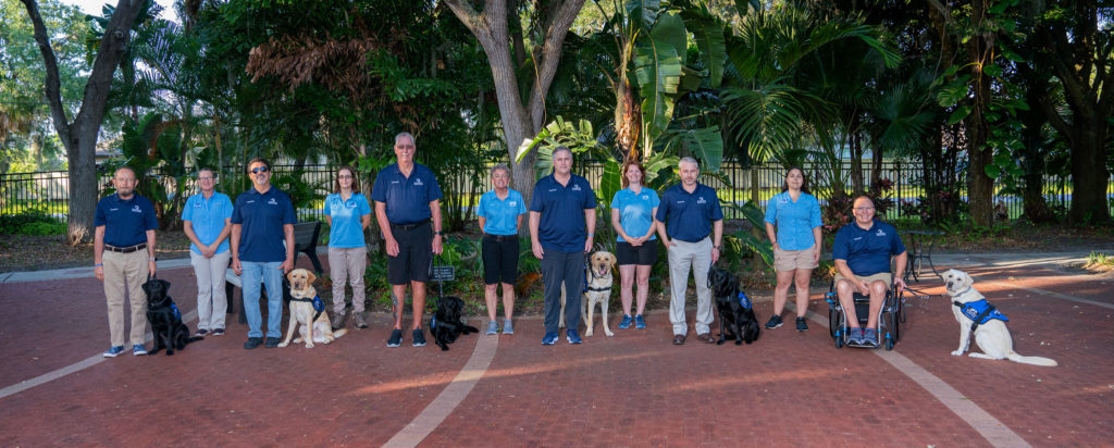 Six graduates are pictured with their service dogs and five trainers stand staggered behind the graduates