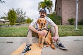 Man sits on curb with his guide dog sitting between his legs