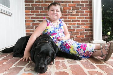 A young child sits on the ground with her arm draped across a black lab