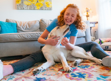 Young woman sits on the ground with yellow lab between her legs and laughs