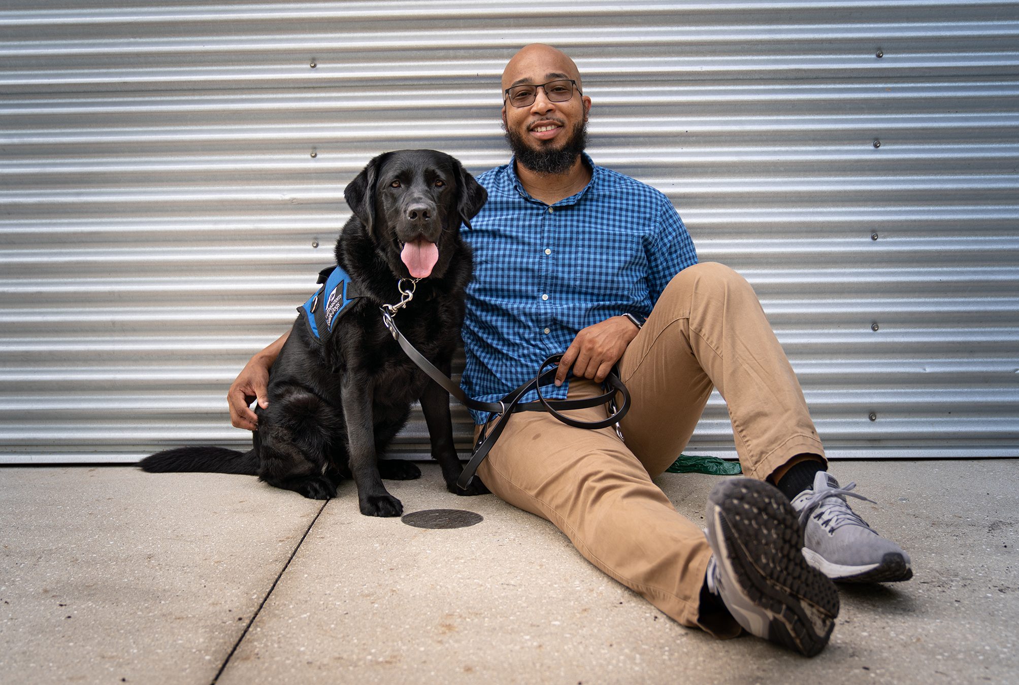 A man sits on the sidewalk with his black lab service dog