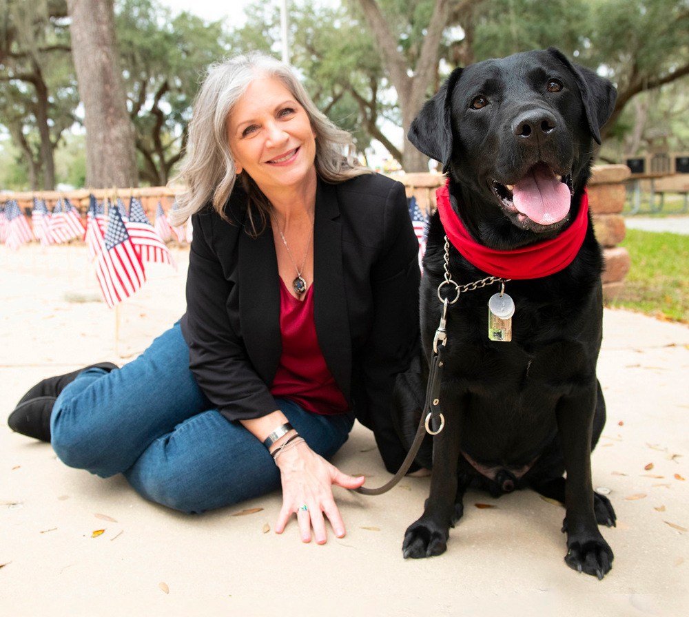 Older woman sits next to black lab who are both smiling and looking at the camera