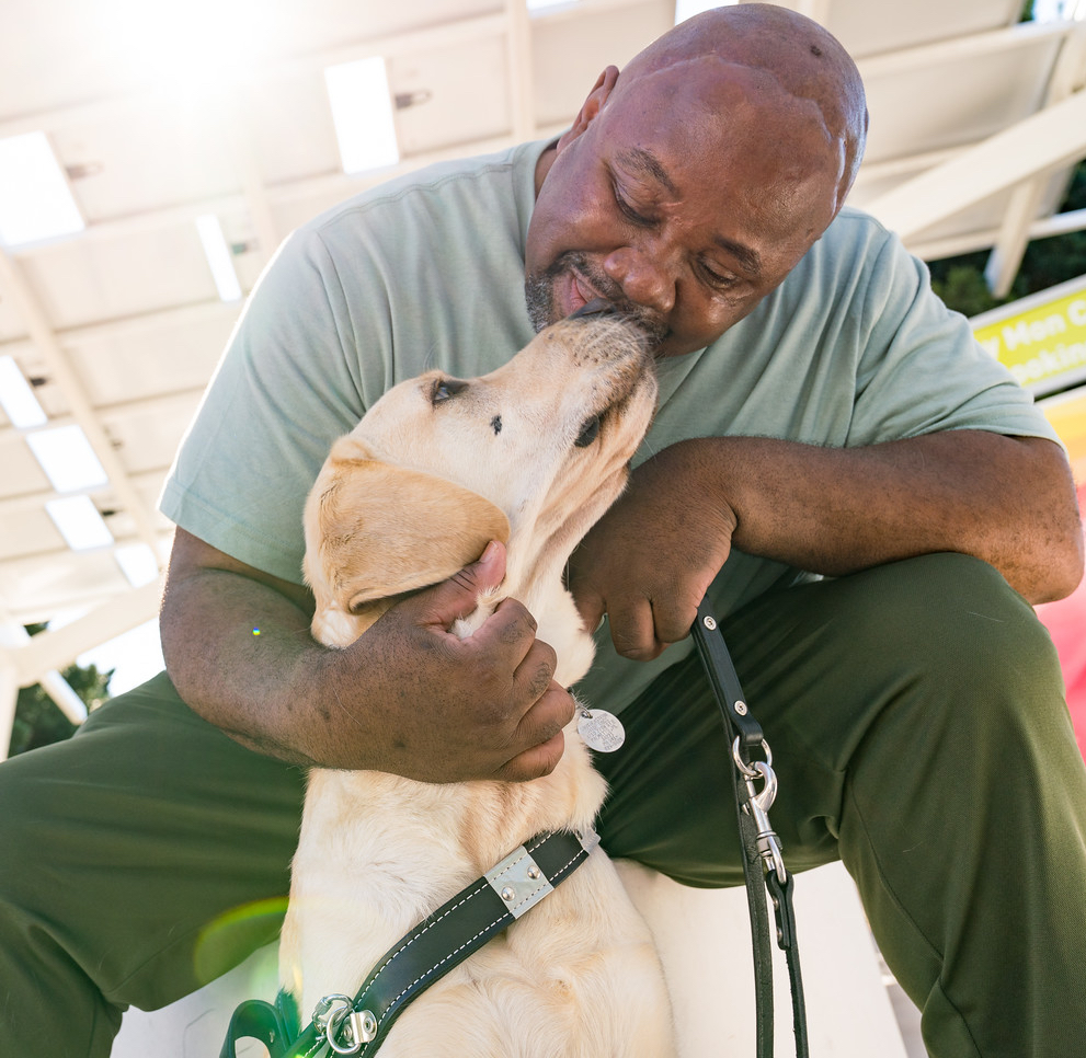 Man is sits with dog between his legs and smiles while the dog is looking up at the man and his nose is touching the mans face