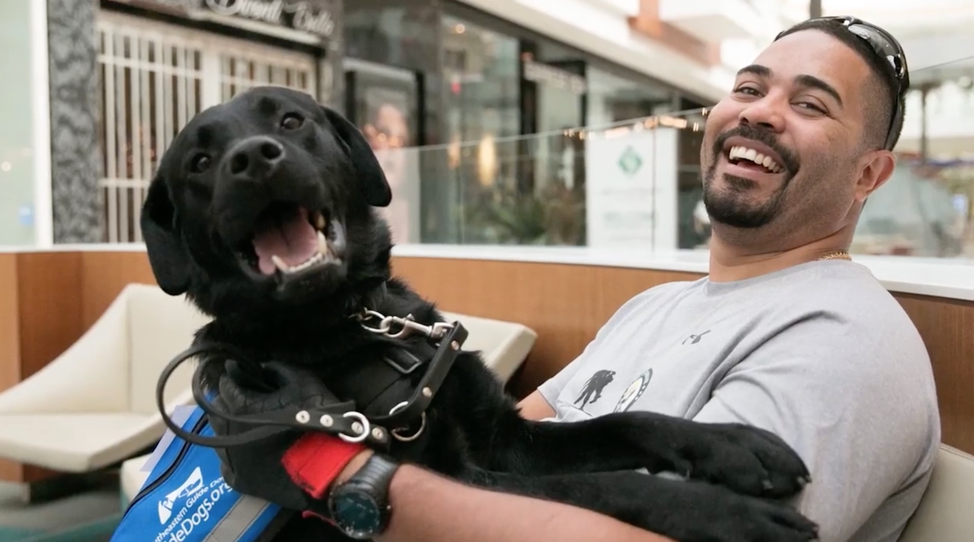 Man sits in chair with a black dogs two front legs on his arm both smiling at the camera
