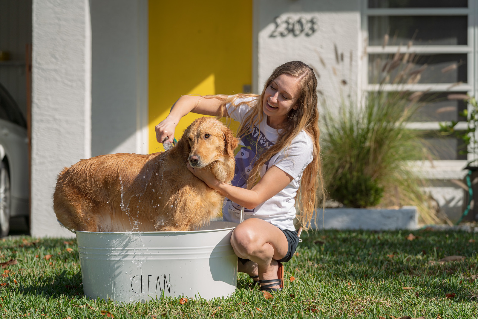 Girl squats down next to dog while she gives a bath to dog in a bucket that says "Clean"