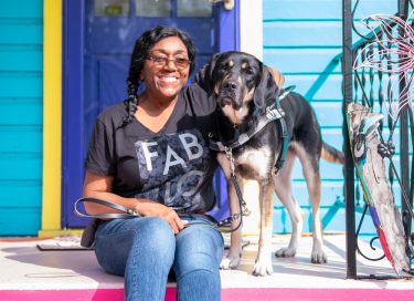 Lady sits on steps and smiles at the camera while her black and an guide dog in harness stands facing the camera