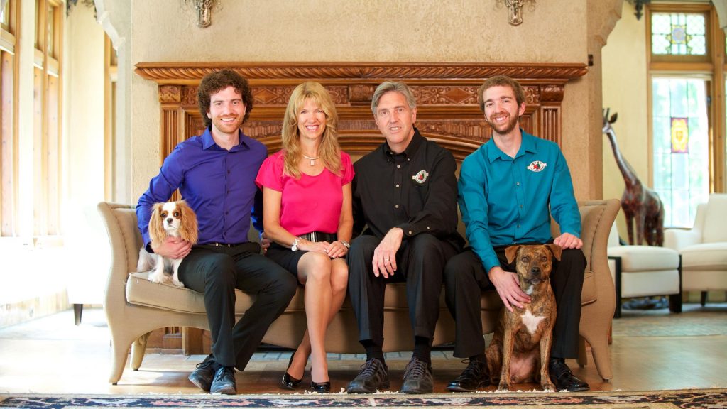 Fromm family sits on couch smiling at the camera with a small dog sitting on the left side of the couch and another dog sitting on the right between a mans kegs