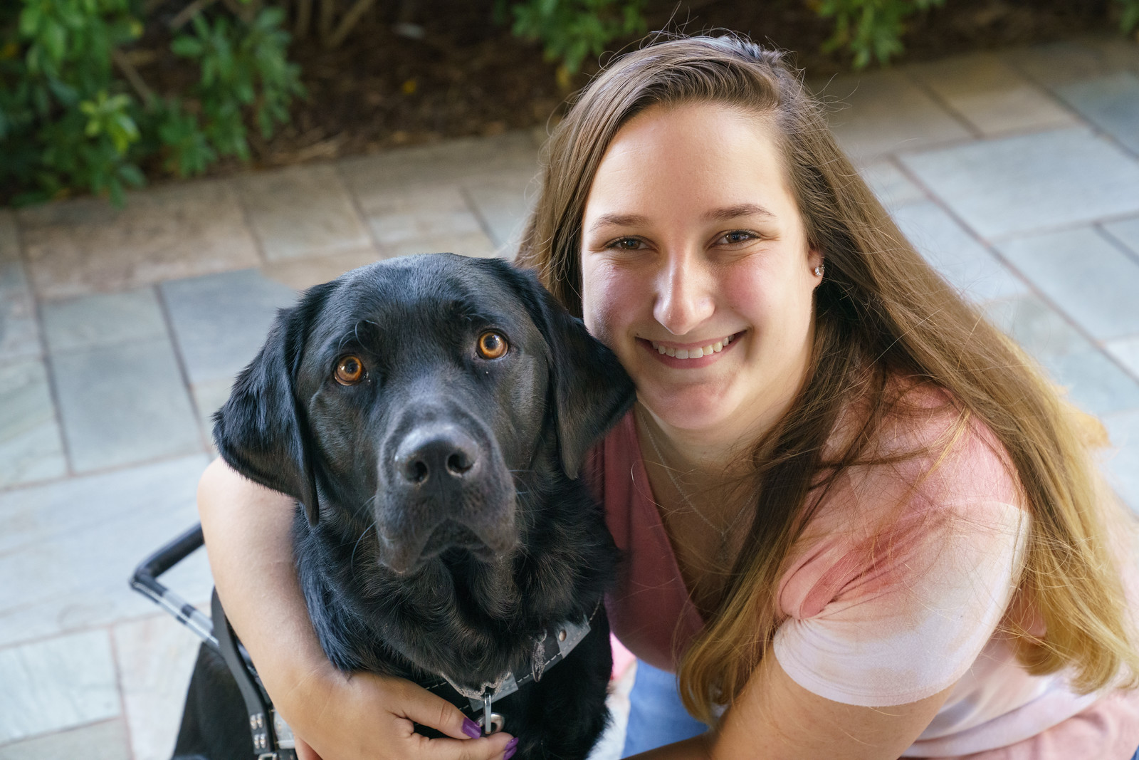 Young woman smiles at the camera while she hugs black lab in harness