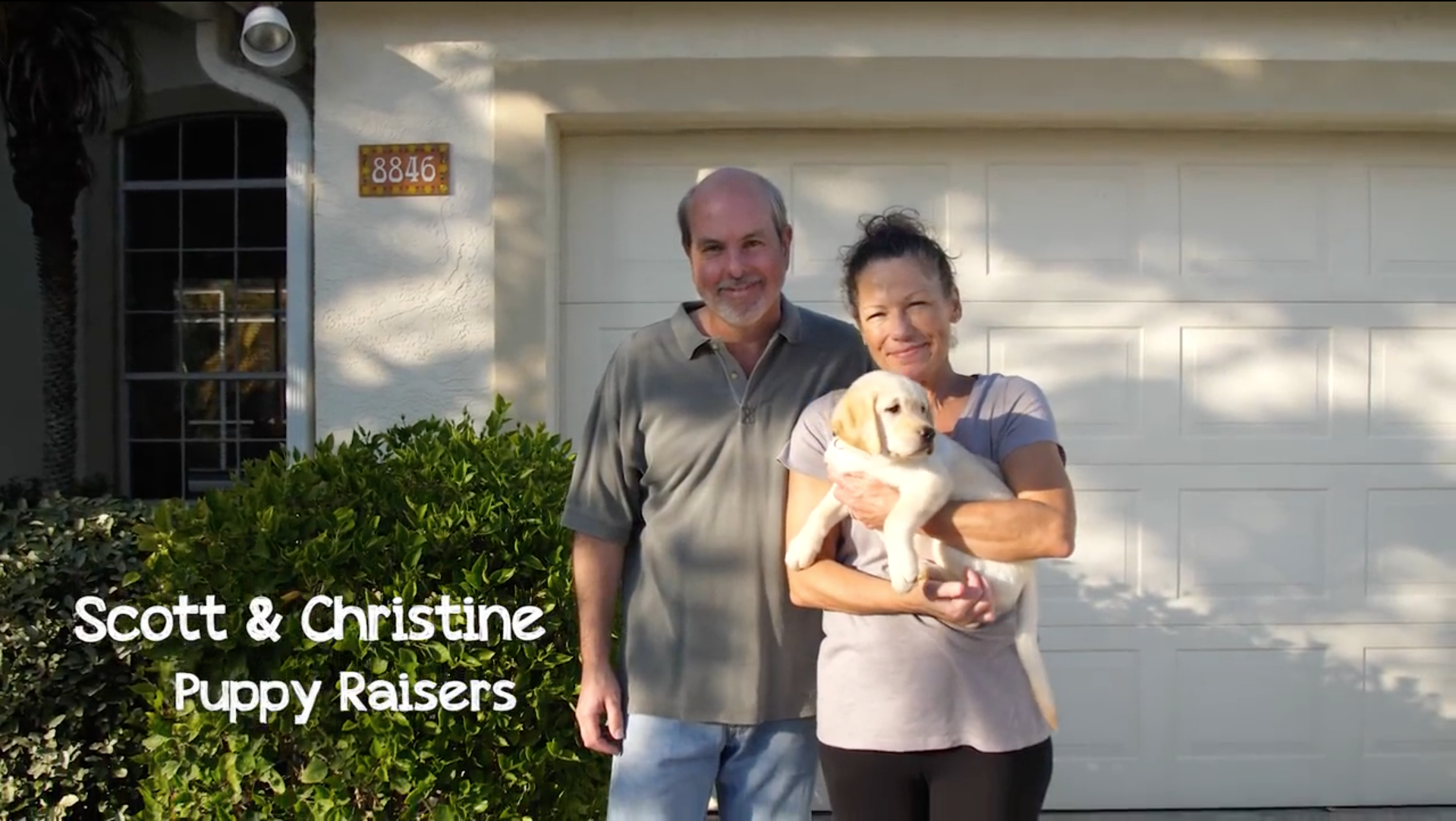 A man and woman stand and smile while she holds a yellow lab puppy that is looking off into the distance