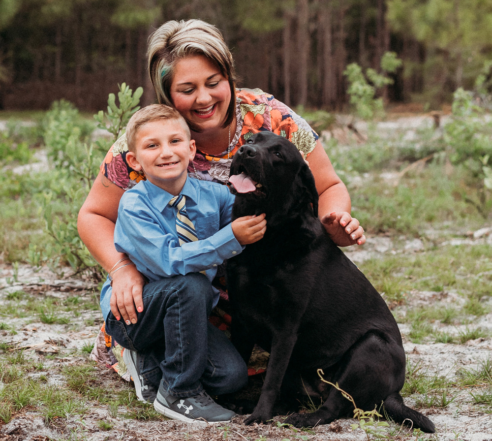 A woman and boy smile while they kneel next to black lab