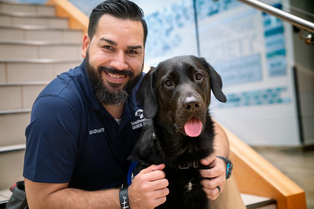 Man sits on stairs with black lab sitting in front of him and both look at the camera and smile