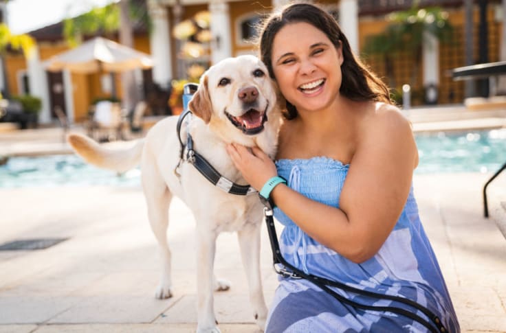Lady sits and smiles with her yellow lab guide dog standing next to her