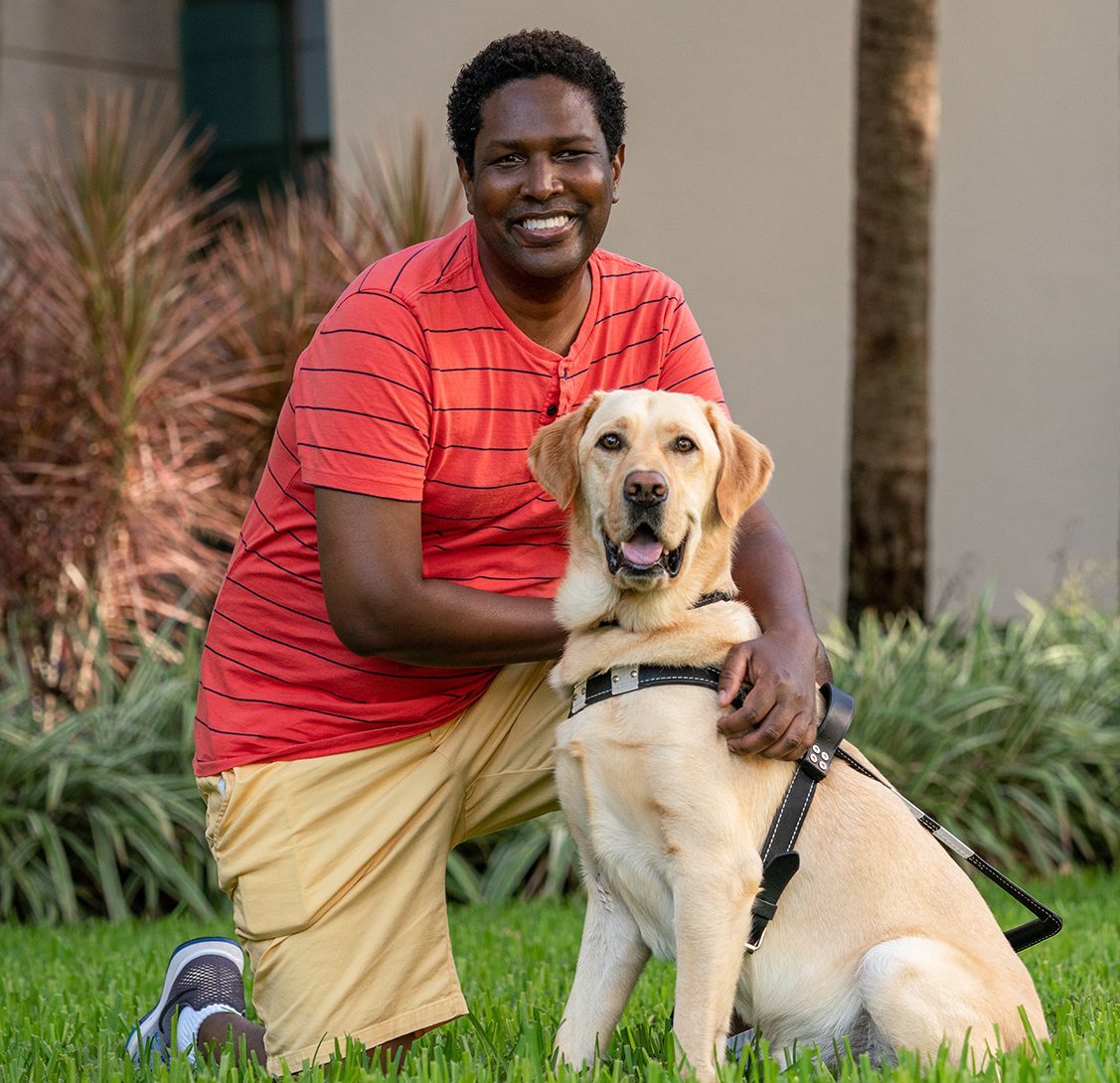 Young man kneels next to yellow goldador in harness who sits both smiling at camera