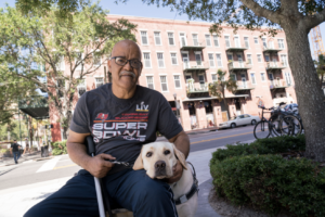A man sits holding walking cane in one hand and pets his guide dog with his other