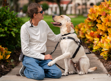 Man kneels next to yellow guide dog whose paw is on his leg