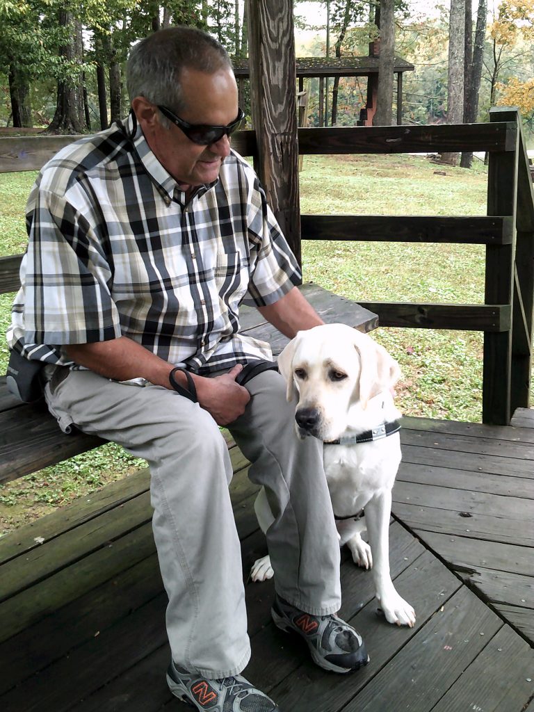 greg sits on a park bench petting his guide dog