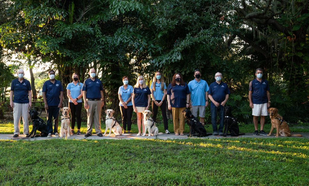 Seven graduates stand with their new guide dogs on the Southeastern Guide Dog campus wearing masks. The four trainers stand behind the graduates, also wearing masks.