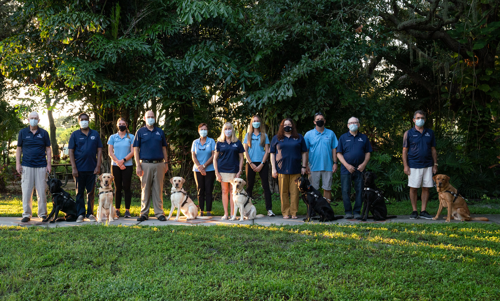 Seven graduates stand with their new guide dogs on the Southeastern Guide Dog campus wearing masks. The four trainers stand behind the graduates, also wearing masks. 