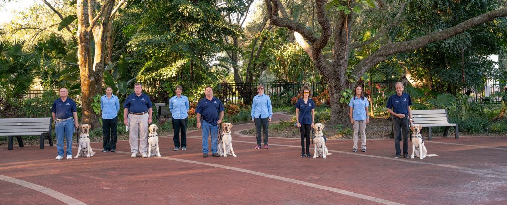 five students stand in a brick courtyard with their new guide dogs on their left sides. The trainers stand in the background.