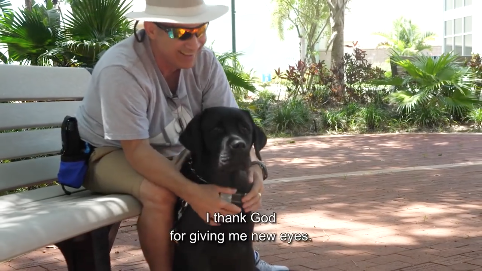 Text: I thank God for giving me new eyes, man sits on a bench and hugs his black lab guide dog