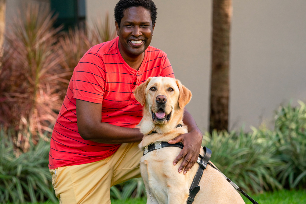 Man kneels on one knee next to yellow goldador guide dog in which both look and smile at the camera