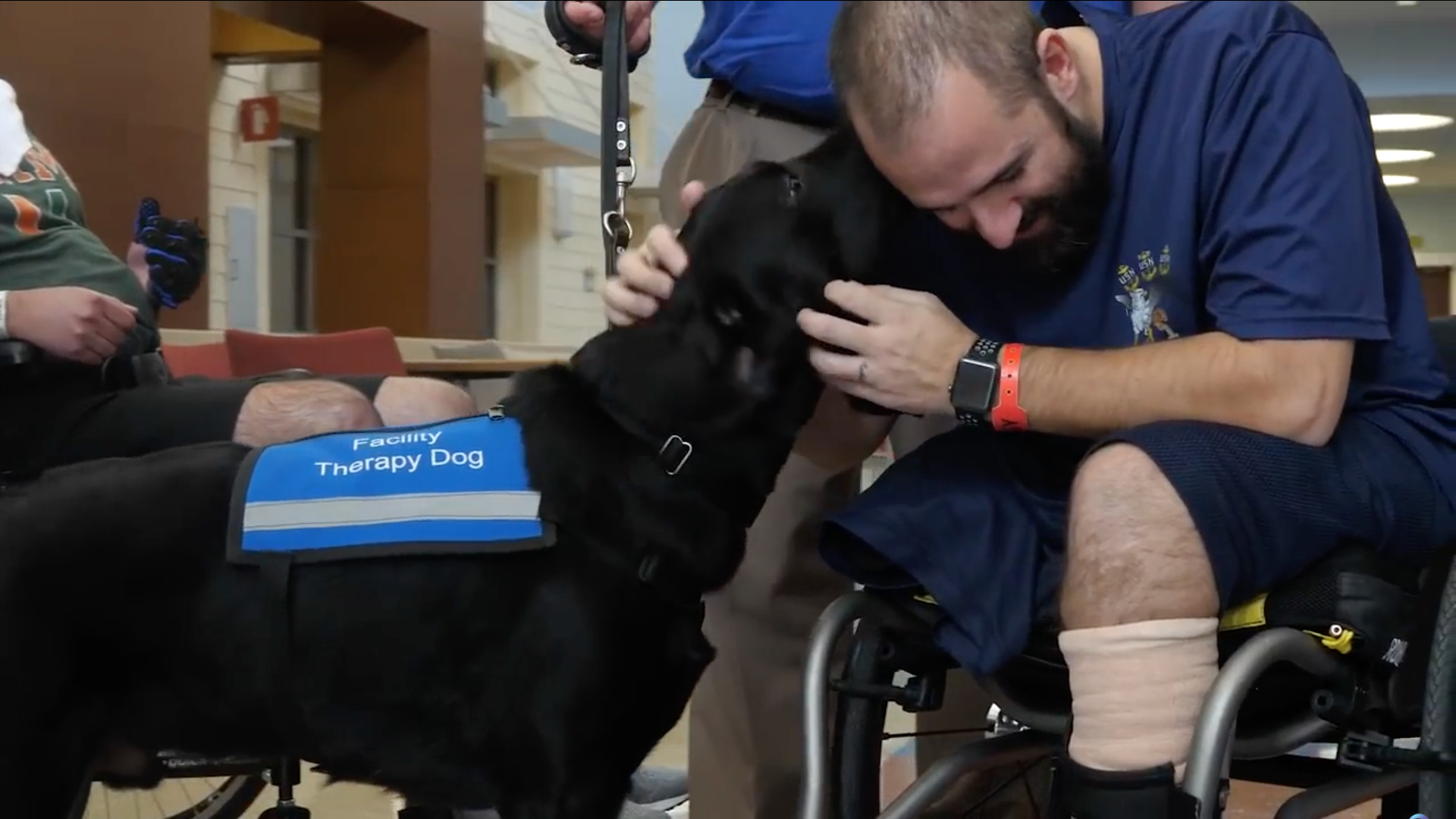 Man in wheelchair leans over and is kissed by a black lab facility therapy dog