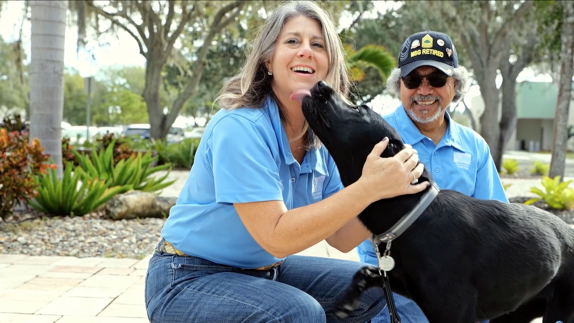 Two puppy raisers sit and see black lab for first time since going in for training, a lady smiles as she is being kissed while the man smiles looking at the two