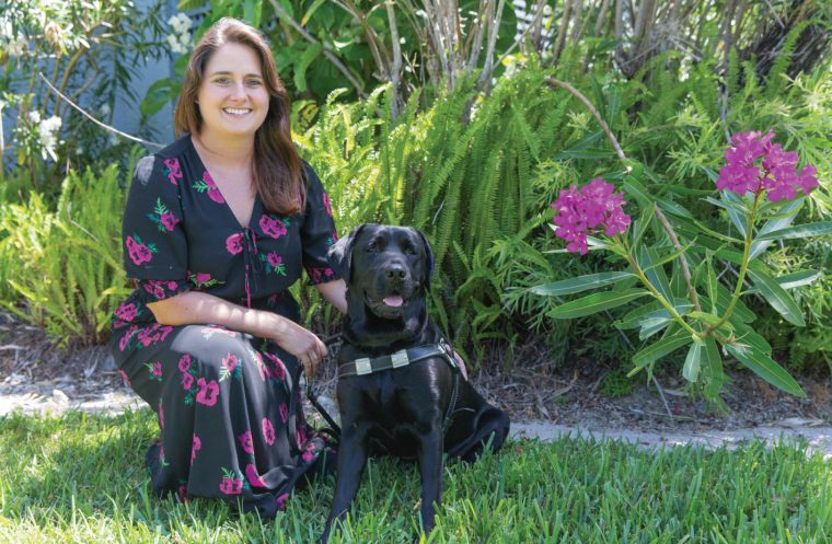 Woman kneels, smiles, and looks at the camera with black lab guide dog sitting next to her smiling