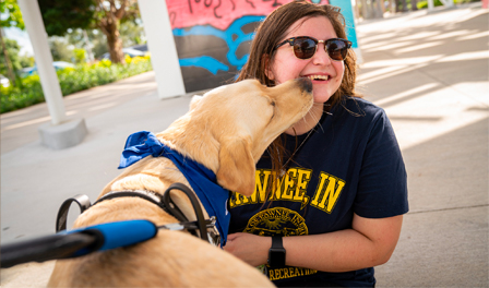 Young adult woman smiles while she is being kissed on the cheek by yellow lab guide dog