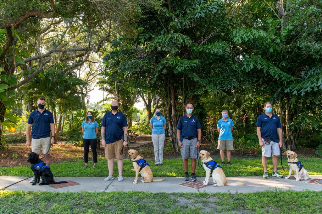 Four veterans stand with their new service dogs, three trainers stand behind them.