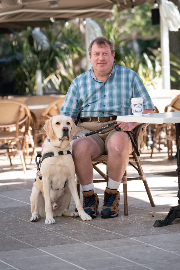 Steven sits at a cafe table with his guide dog sitting next to him