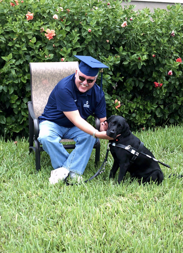 Troy sits in a lawn chair wearing a graduation cap holding his dogs head