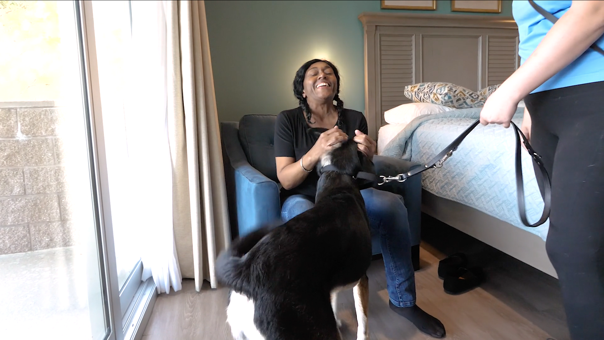 Woman sits in chair and pets black labs head and she smiles widely