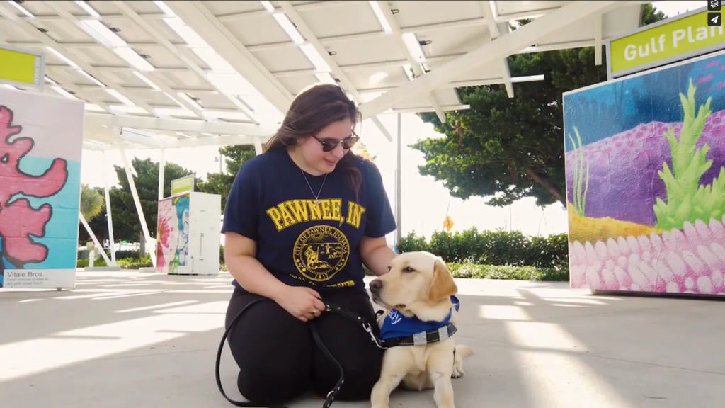 Woman sits and smiles down at yellow guide dog as she pets the top of the dog