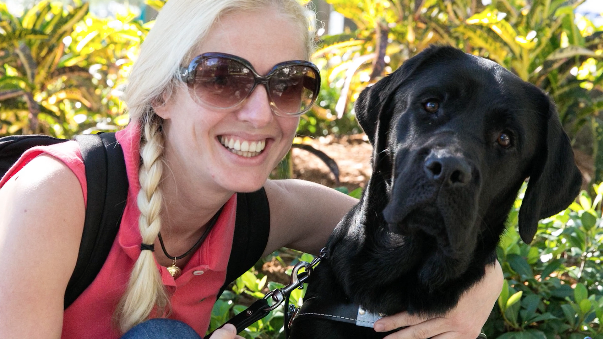 Woman leans over and has arm around black guide dog while she smiles at the camera