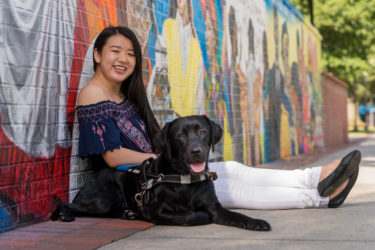 A young Asian girl leans against painted wall with black Lab guide dog