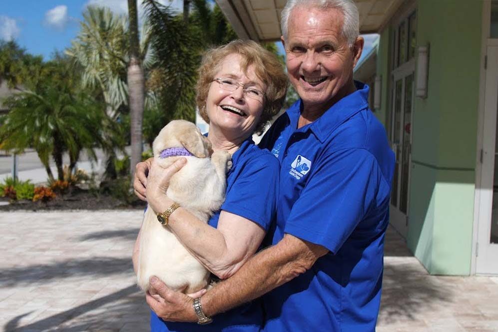 An older couple smiling at the camera while they hold a yellow lab puppy.