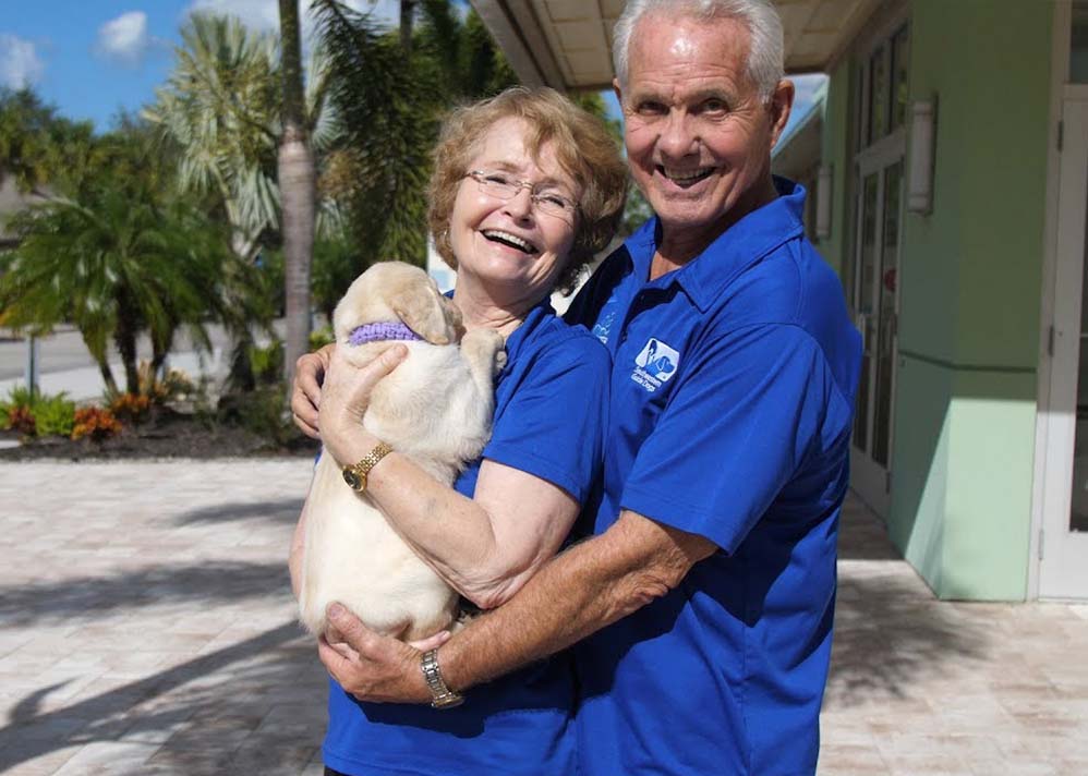 An older couple smiling at the camera while they hold a yellow lab puppy.