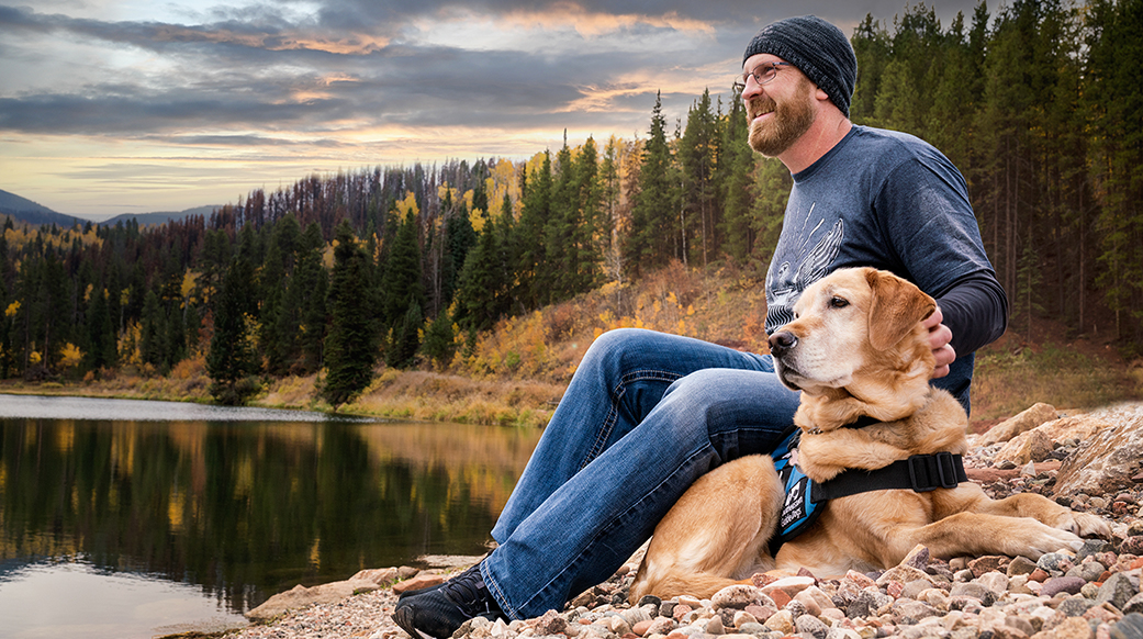 A man sits on a rocky beach surrounded by forest with his yellow lab service dog. They both look off into the distance.