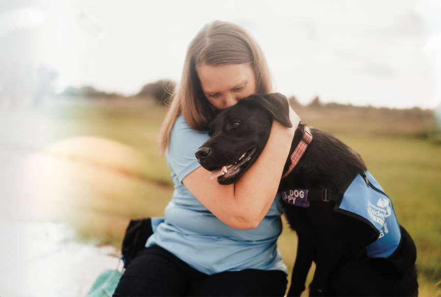 A woman hugging black Lab puppy in a blue coat