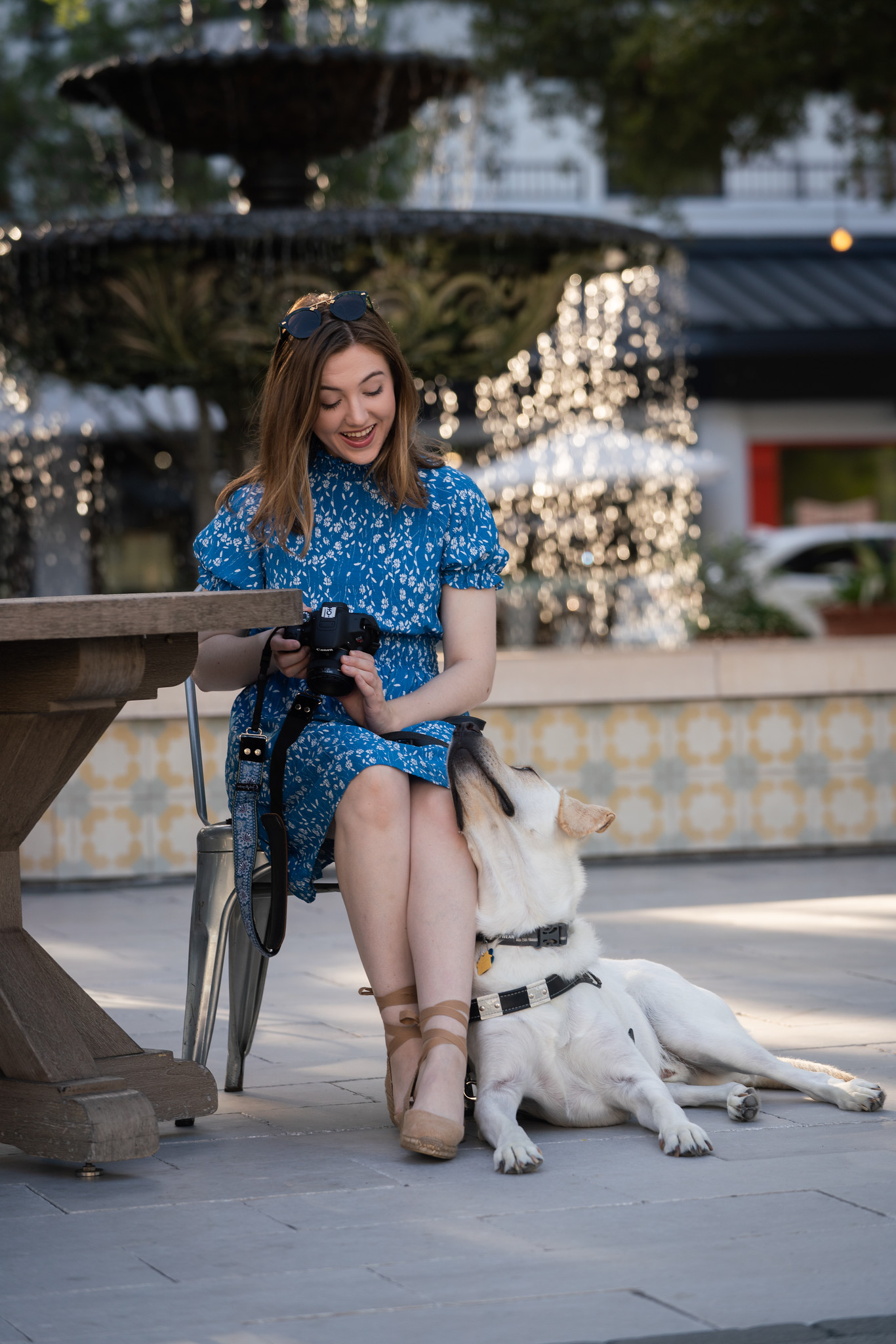 A young woman in a blue dress sits outside at a patio table near a fountain with her yellow lab guide dog laying next to her chair