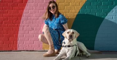 Clare Weigel and guide dog Fin posing on a sidewalk in front of a rainbow-striped wall.