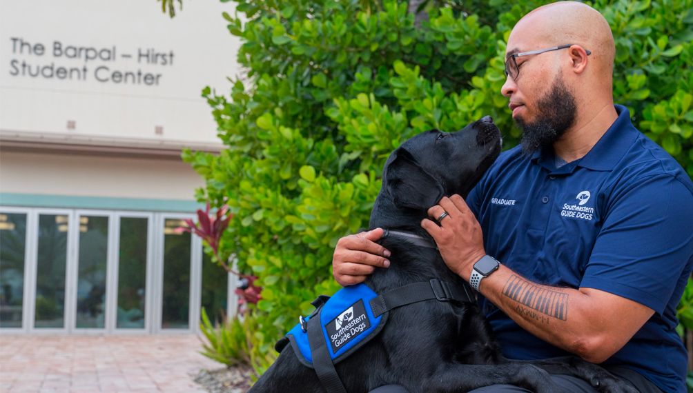 A black lab service dog stands with his front two legs over his veterans lap and kisses him on the chin