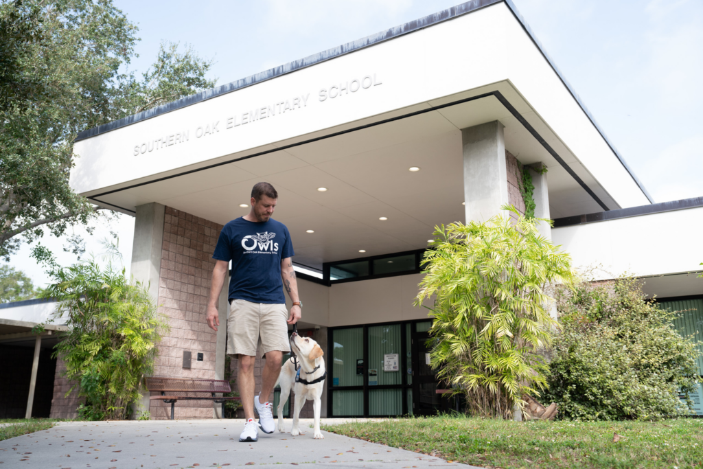 A man walking with his yellow lab service dog outside of an elementary school