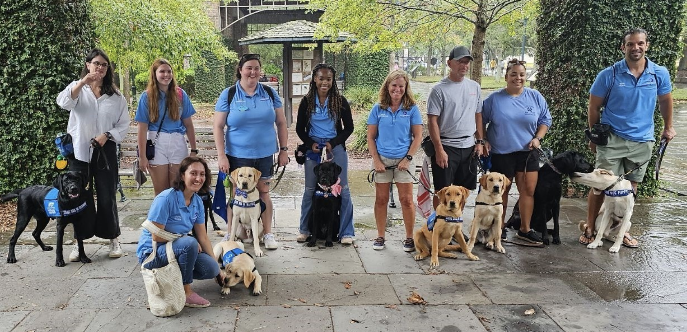 A group of individuals stand for a photo while their dogs in training sit in front of them.